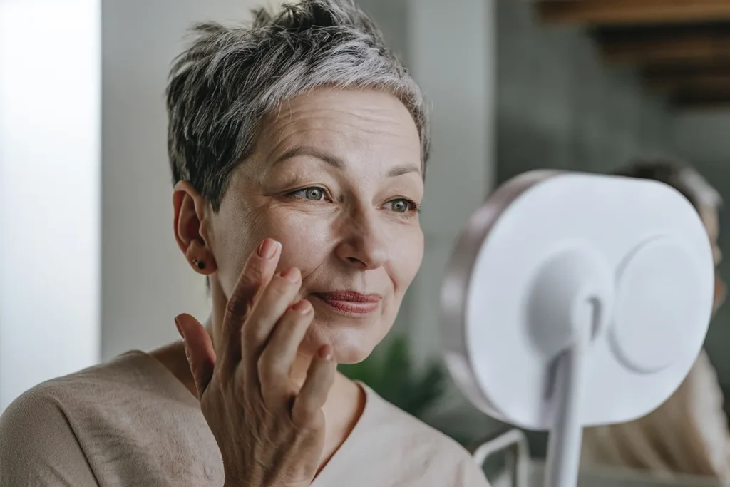 Older woman with short gray hair examining her face in a mirror, gently touching her cheek while standing in a well-lit bathroom.