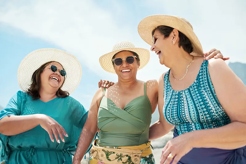 Three older women in sun hats and beachwear laughing and enjoying a sunny day at the beach.