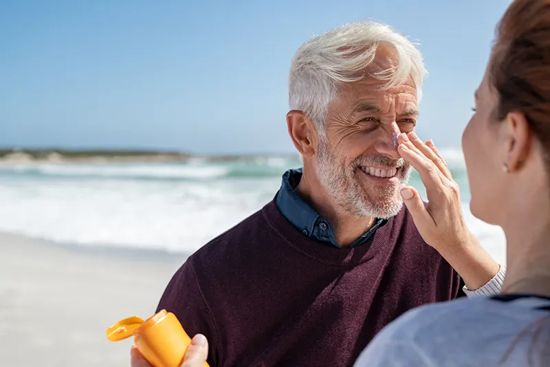 Smiling older man having sunscreen applied to his face by a woman at the beach, with the ocean in the background.