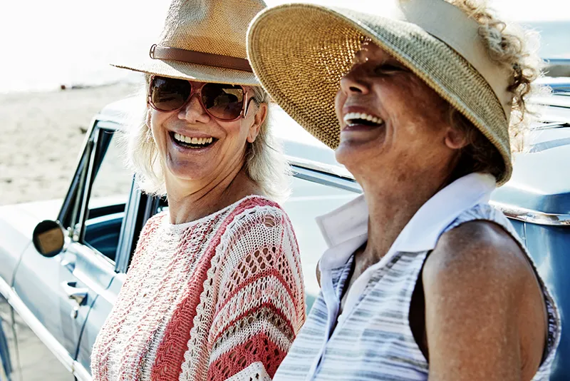 Two older women wearing sun hats and sunglasses smiling and laughing beside a classic car at the beach.
