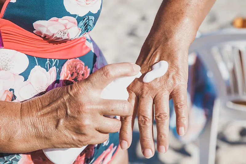 Close-up of a person applying sunscreen to the back of their hand while wearing a floral swimsuit at the beach.