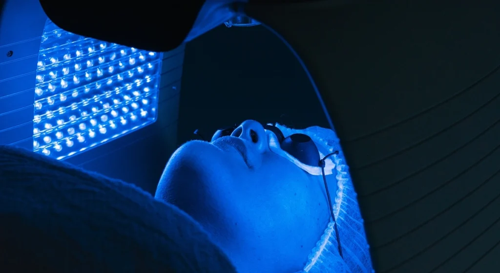 Patient undergoing blue light therapy (photodynamic therapy) for skin treatment, lying under a specialized lamp with protective eyewear.