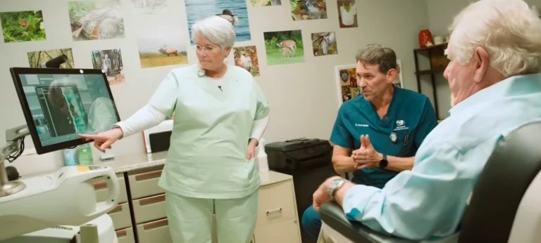 Medical consultation scene where a clinician points to imaging displayed on a monitor while discussing results with two colleagues and an older patient seated in an exam chair inside a clinical treatment room.
