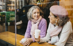 Two older women smiling and chatting while holding coffee cups at a café table, representing an active, relaxed lifestyle after skin cancer care and treatment.