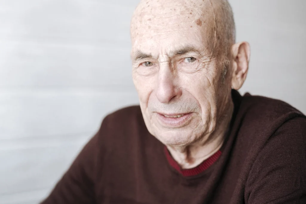Close up portrait of an older man with a shaved head and visible sun spots on his skin, looking calmly toward the camera in a softly lit indoor setting.