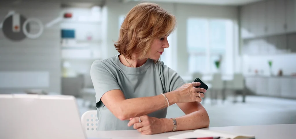 Middle aged woman seated indoors examining a spot on her forearm using a smartphone camera, suggesting early skin self check in a bright, modern home setting.