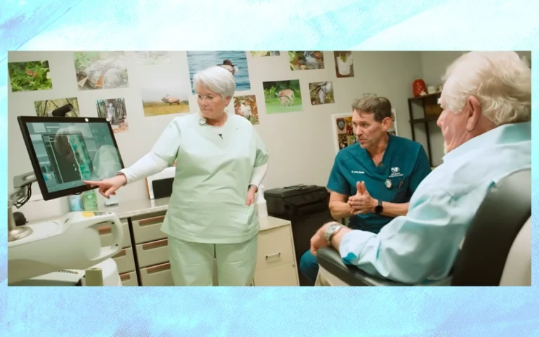 Medical consultation scene where a clinician points to imaging displayed on a monitor while discussing results with two colleagues and an older patient seated in an exam chair inside a clinical treatment room.