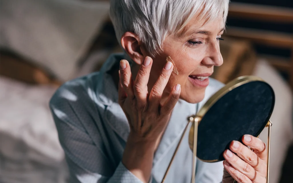 Older woman with short gray hair examining a small spot on her cheek while looking into a handheld mirror in a softly lit indoor setting.