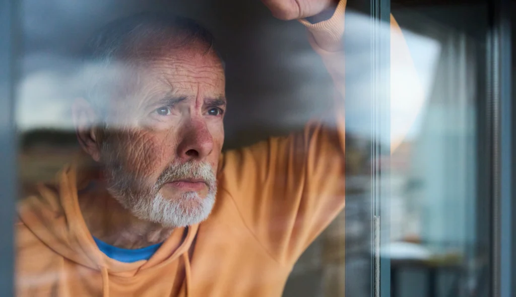 Older man looking through a window while sitting indoors in soft natural light.